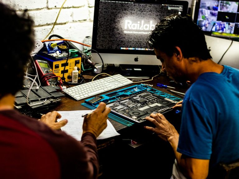 Two individuals working on electronics at a desk, with computer and tools visible.