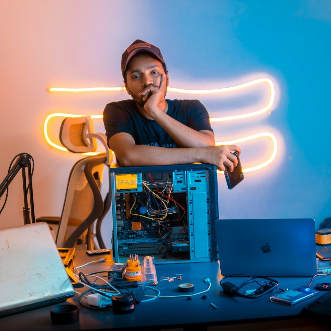 A person sitting with a computer tower, surrounded by various tech gadgets and neon lighting.